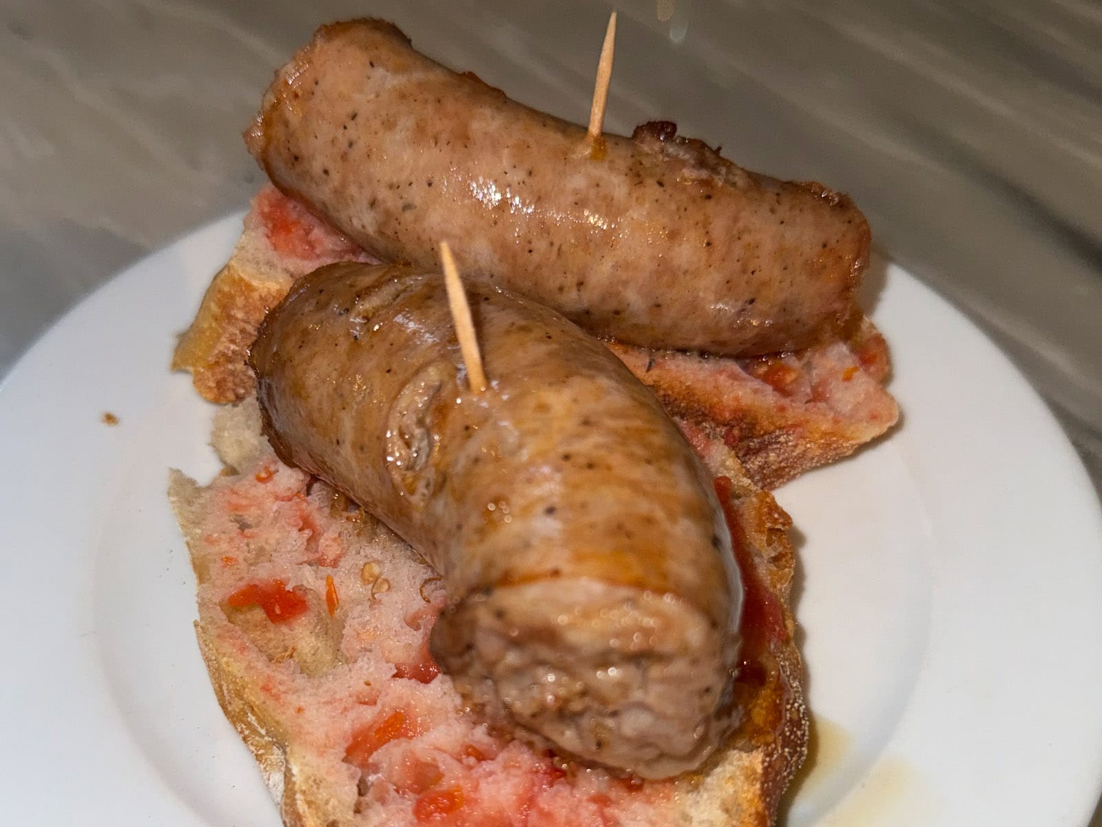 Pincho de butifarra del Bar La Plata: butifarra a la plancha sobre pan con tomate, sujeta con un palillo, en plato blanco