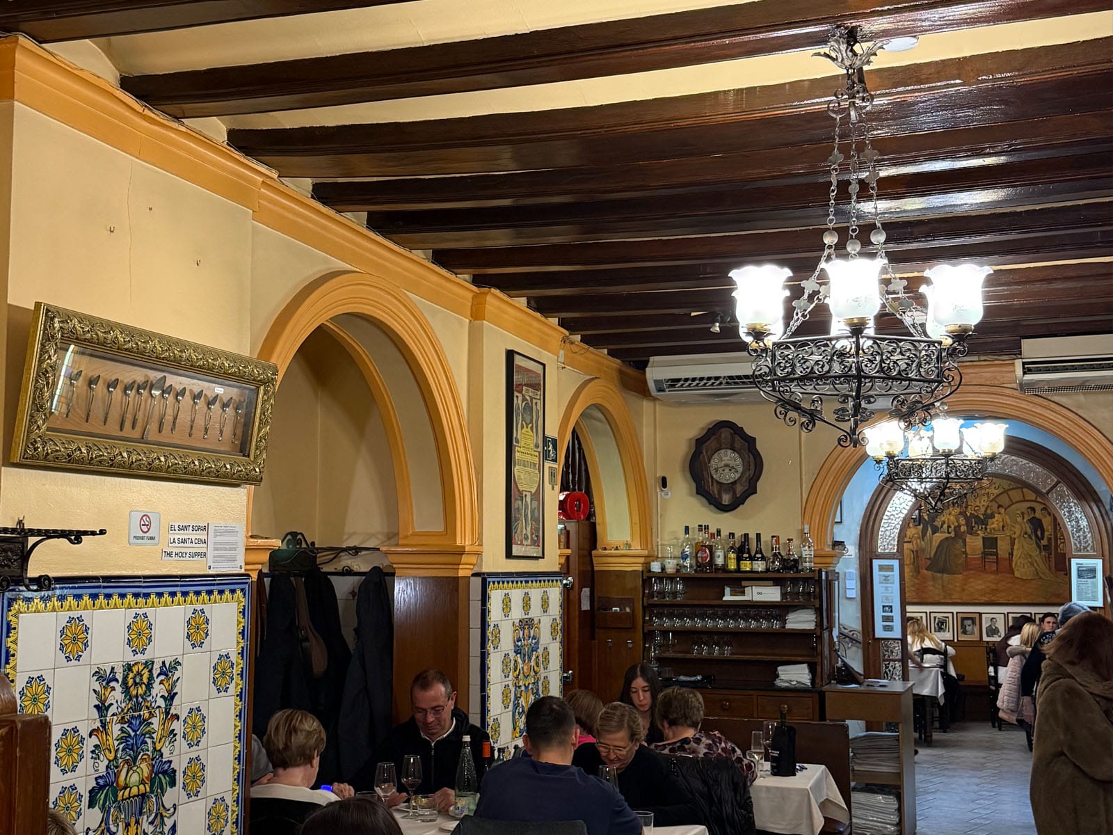Comedor de Can Culleretes: paredes amarillas con azulejos azules, vigas de madera oscura, lámparas de forja y una vitrina con cucharillas antiguas enmarcada en la pared