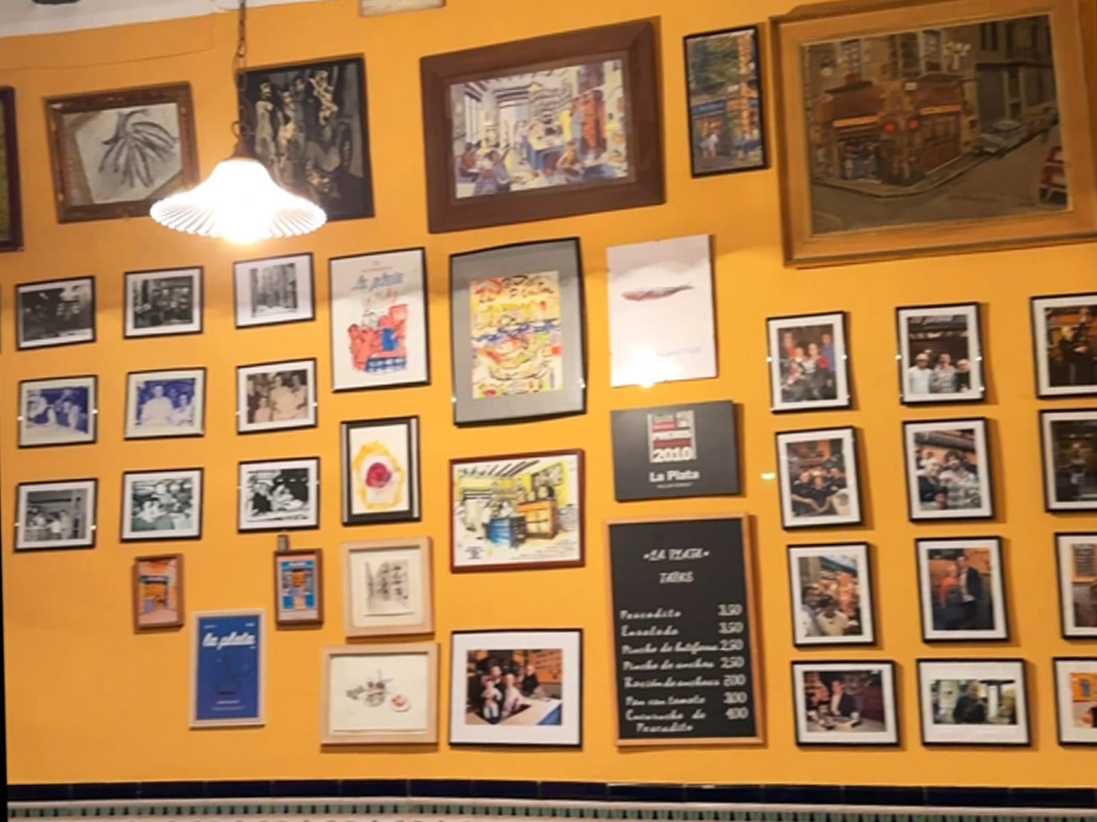 Pared amarilla del Bar La Plata cubierta de cuadros, dibujos y fotografías enmarcadas, con la pizarra de las cuatro tapas y sus precios en el centro