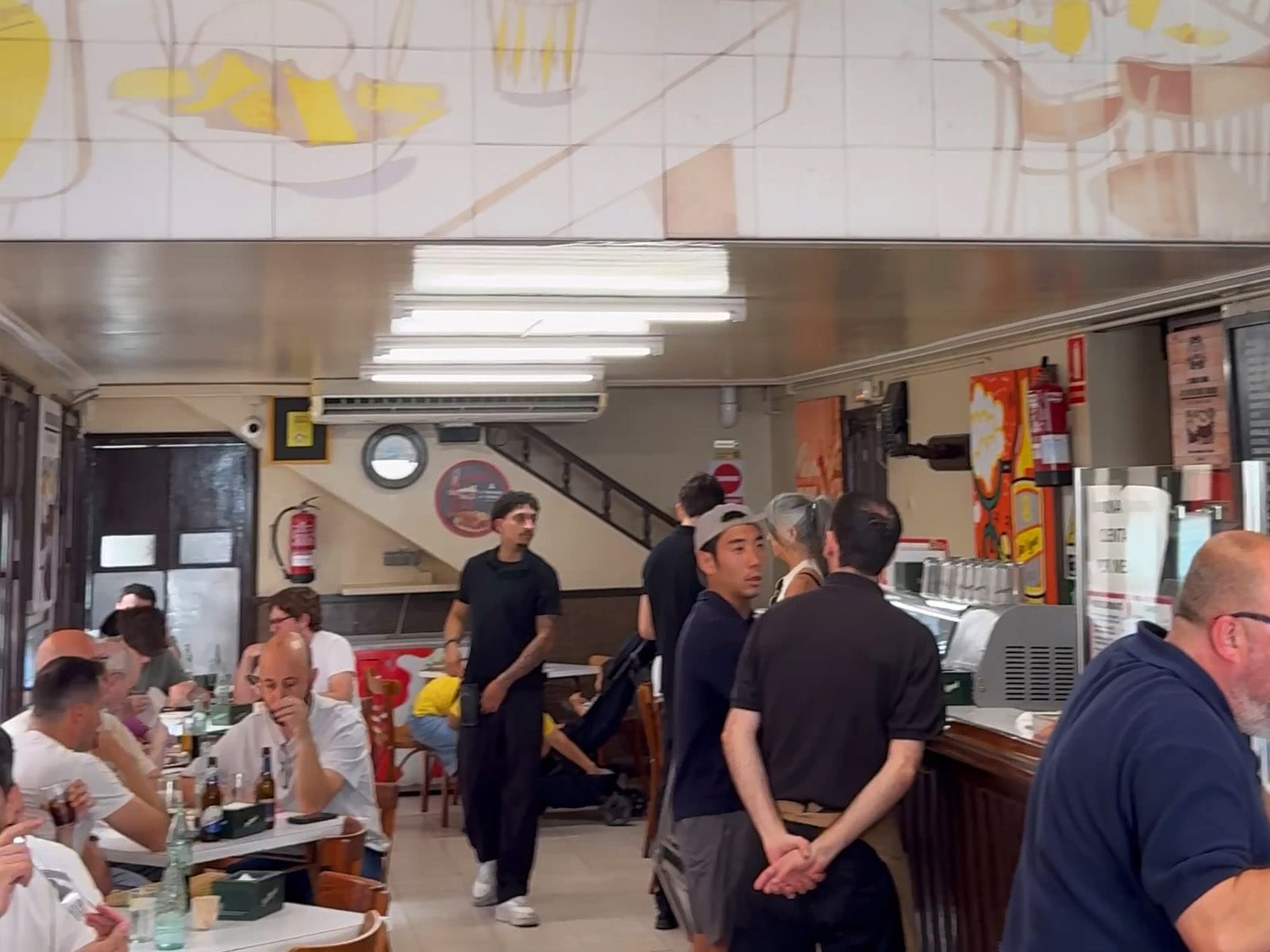 Interior del Bar Tomás de Sarrià lleno a mediodía, con las mesas de mármol blanco, el techo de azulejos pintados y los camareros sirviendo entre las mesas