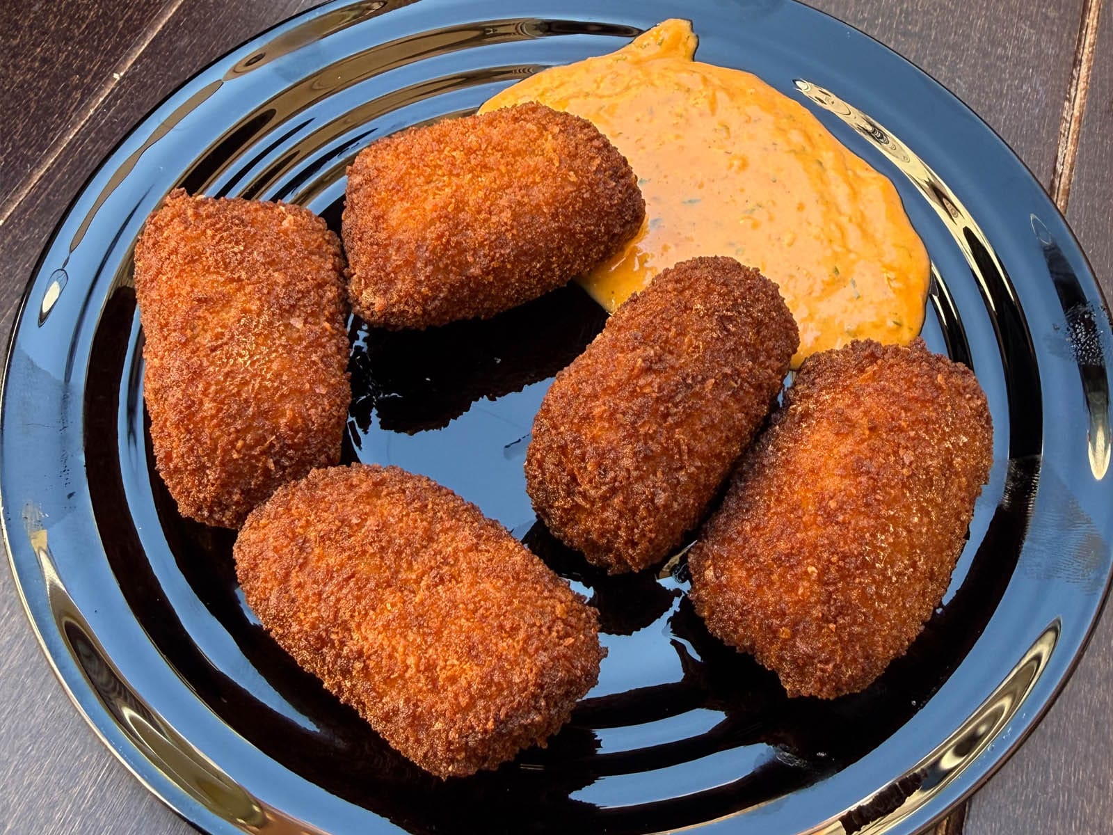 Cinco croquetas de calçots doradas sobre un plato negro con salsa anaranjada