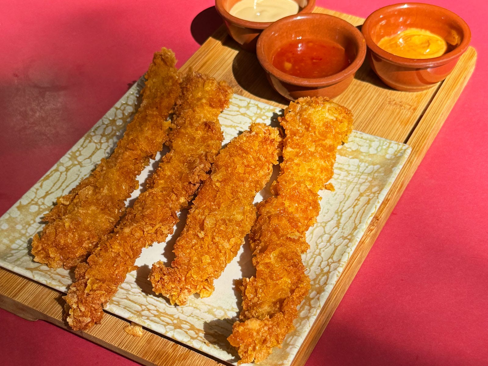 Delicias de pollo del Canut: cuatro tiras de pollo con rebozado muy crujiente sobre tabla de madera, con tres cuencos de barro con salsas orientales