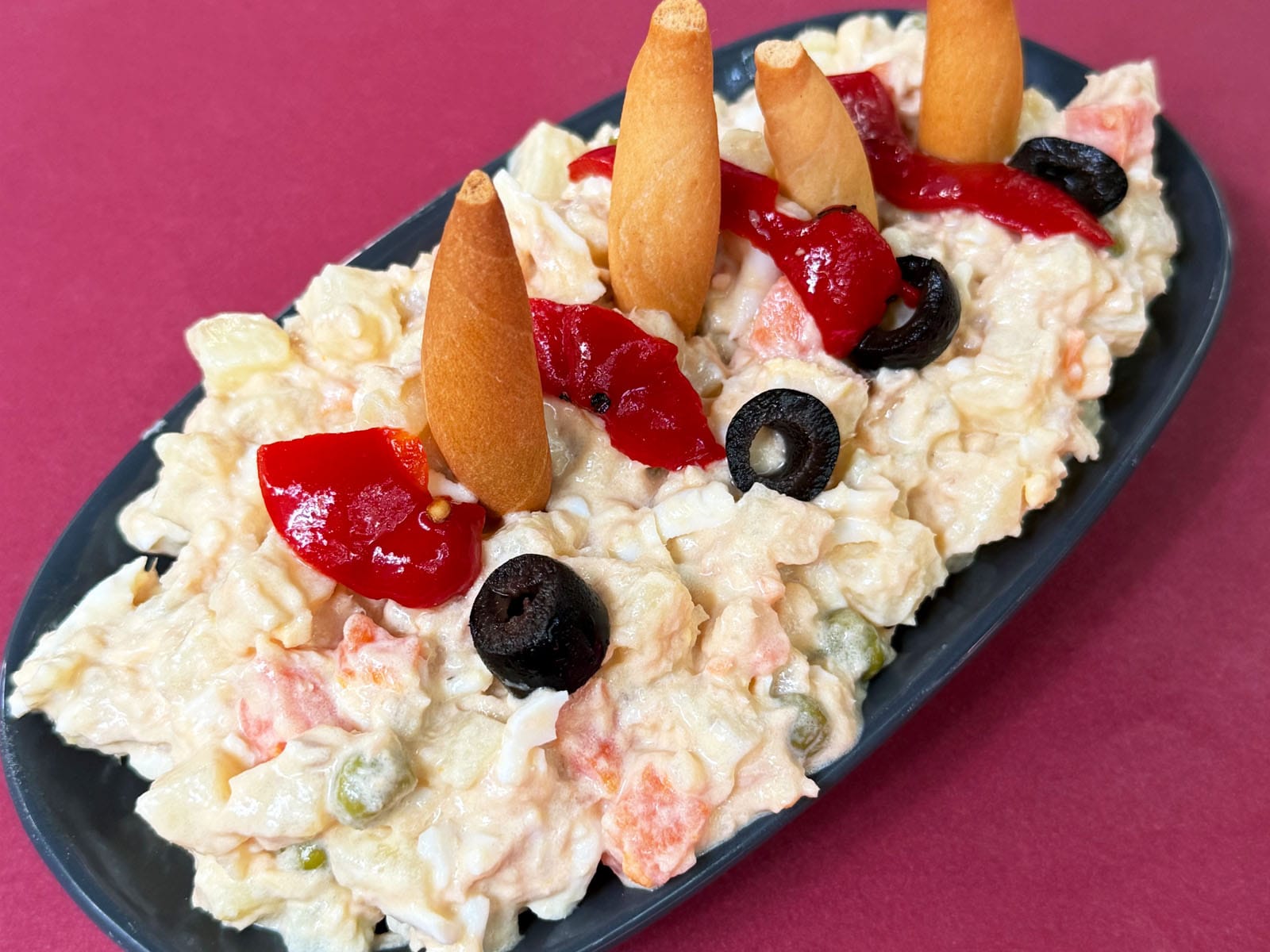 Ensaladilla del Canut servida en fuente negra: patata, huevo, guisantes y zanahoria ligados con mayonesa, coronada con tiras de pimiento del piquillo, aceitunas negras y picos de pan