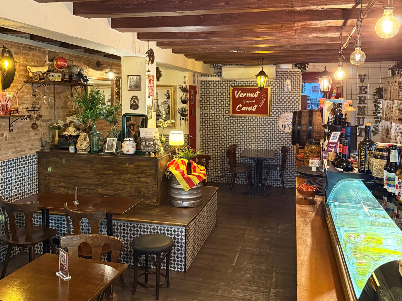 Interior de Vermut i Força al Canut: paredes de ladrillo visto, vigas de madera, mosaico hidráulico, el cartel rojo con el nombre del local y la vitrina de encurtidos en la barra